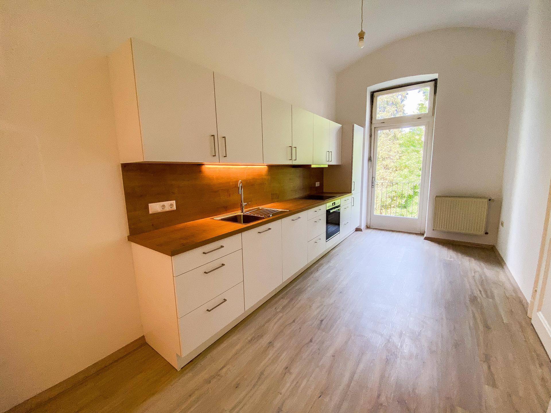 Helle Küche mit weißer Einbauküche, Holzdekor und Blick ins Grüne durch die Balkontür.