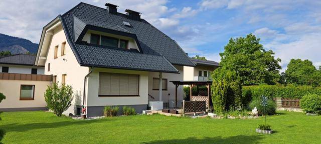 Seitenansicht des Hauses mit großem Garten, Terrasse und Bergblick unter bewölktem Himmel.