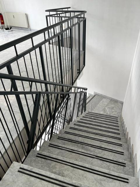 Geschwungene Marmortreppe mit schwarzem Metallgeländer und rutschfesten Streifen, die nach unten führt.