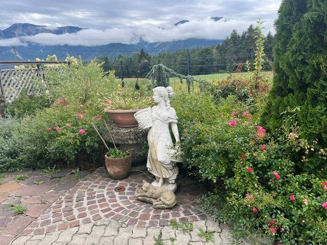 Romantischer Gartenbereich mit Steinpflaster, Blumenbeeten und einer klassischen Statue vor Bergkulisse.