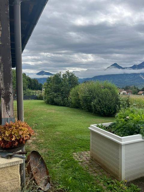 Blick von einer überdachten Terrasse auf einen weitläufigen Garten mit Rasen und Bergpanorama.