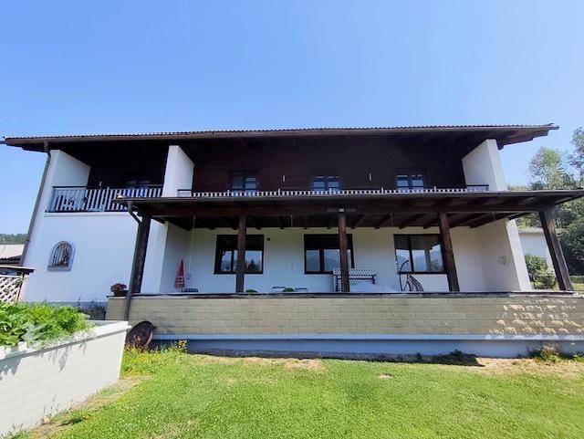 Rückseite des Hauses mit überdachter Terrasse und Balkonen, umgeben von grüner Landschaft.