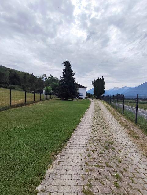 Lange gepflasterte Auffahrt, die zum Haus führt, umgeben von weitläufigen Grünflächen und Bergen.