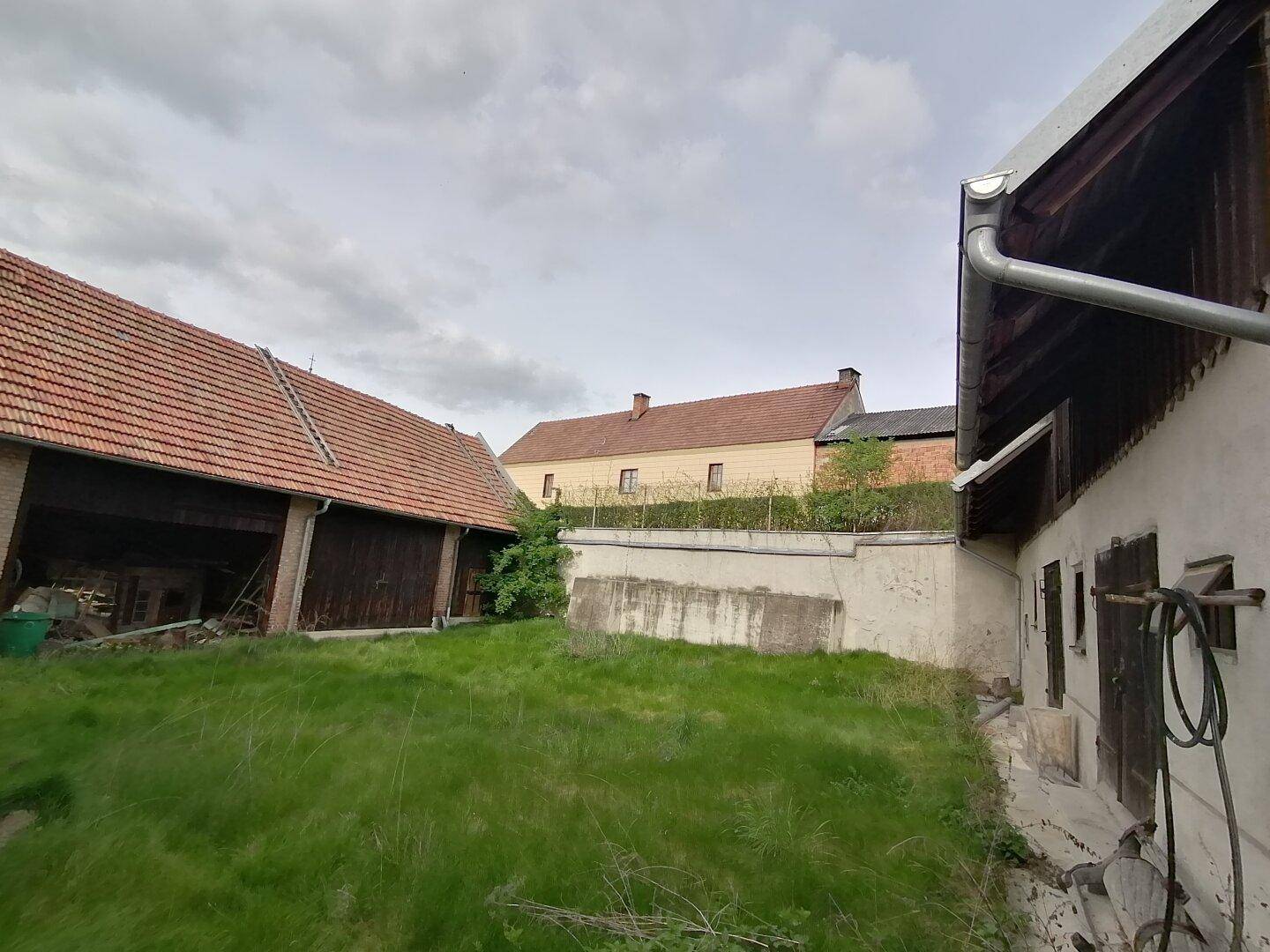 Blick auf die Scheune und ein weiteres Gebäude mit einer Betonmauer im Vordergrund, umgeben von Gras.