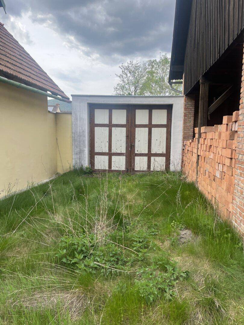 Kleine Garage mit Holztüren, umgeben von hohem Gras und einer Ziegelmauer.