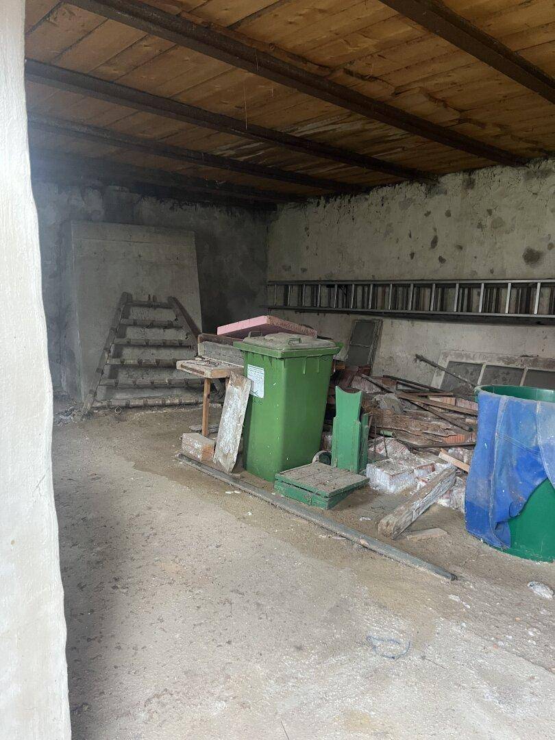Unaufgeräumter Abstellraum mit Betonboden, Holzbalkendecke und verschiedenen gelagerten Gegenständen.