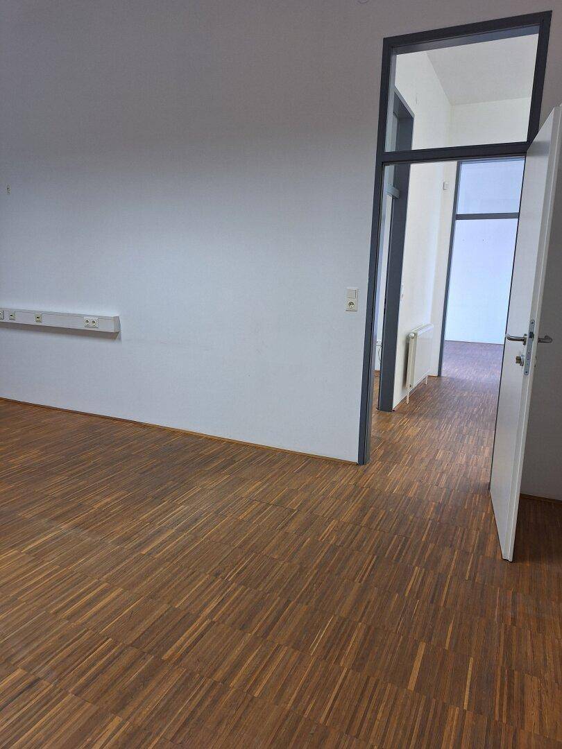 Heller Büroraum mit Holzboden und Zugang zu einem Flur durch eine Glastür.