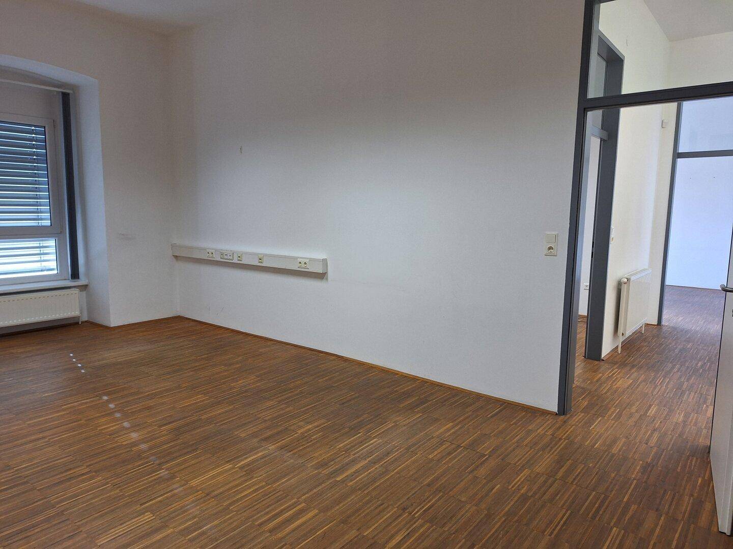 Geräumiger Büroraum mit Holzboden, Fenster mit Jalousien und Steckdosenleisten an der Wand.