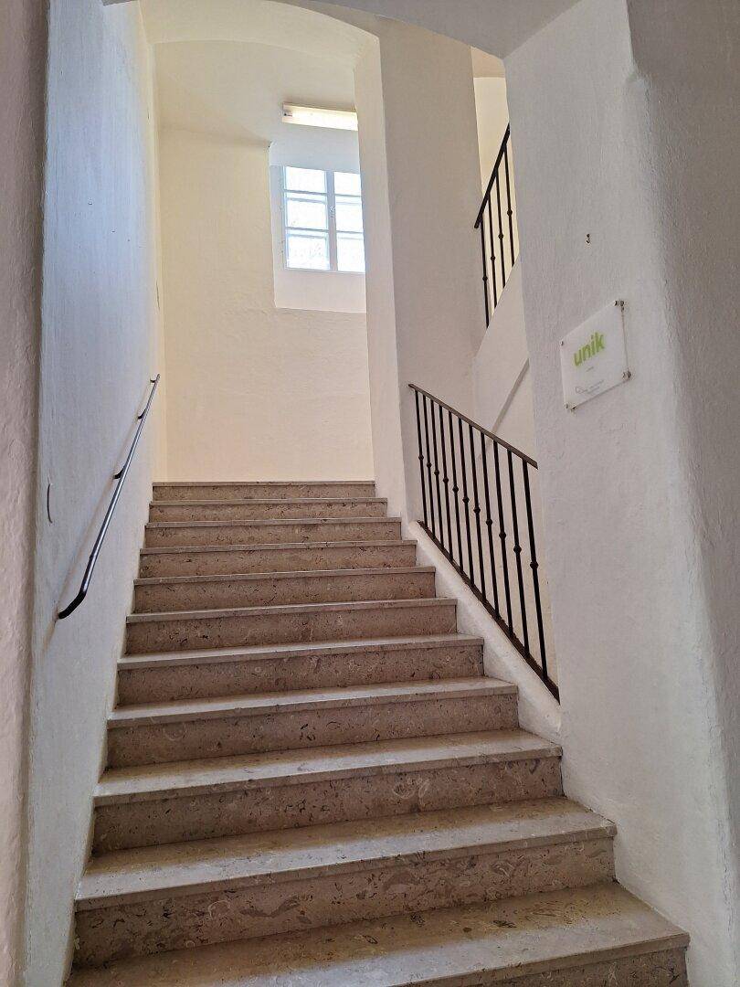 Steintreppe mit Metallgeländer und einem Fenster, das natürliches Licht in das Treppenhaus bringt.