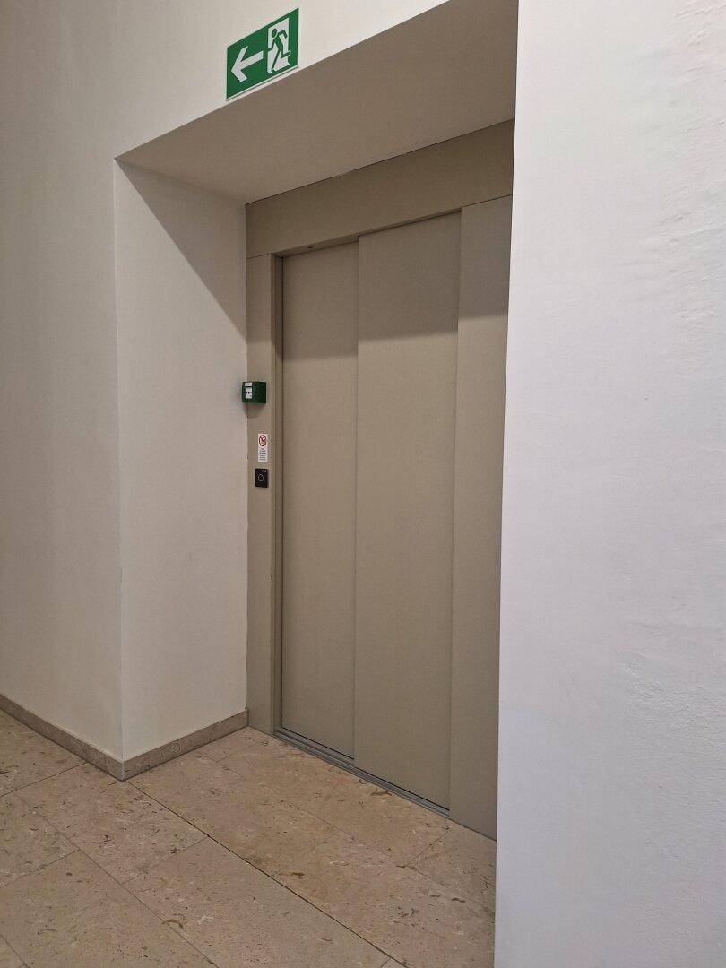 Moderner Aufzug in einem Flur mit hellen Fliesen und einem Notausgangsschild.