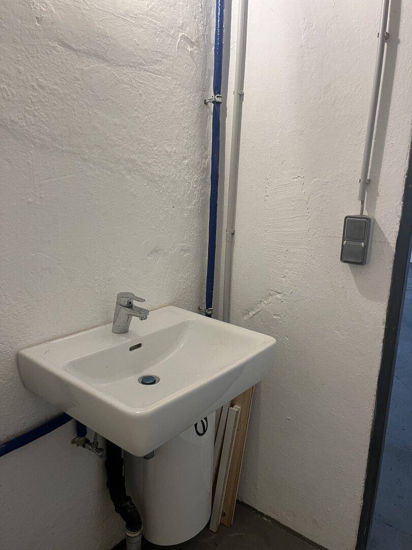 Kleiner Waschbereich mit einem einfachen Waschbecken und sichtbaren blauen Wasserleitungen an der Wand.