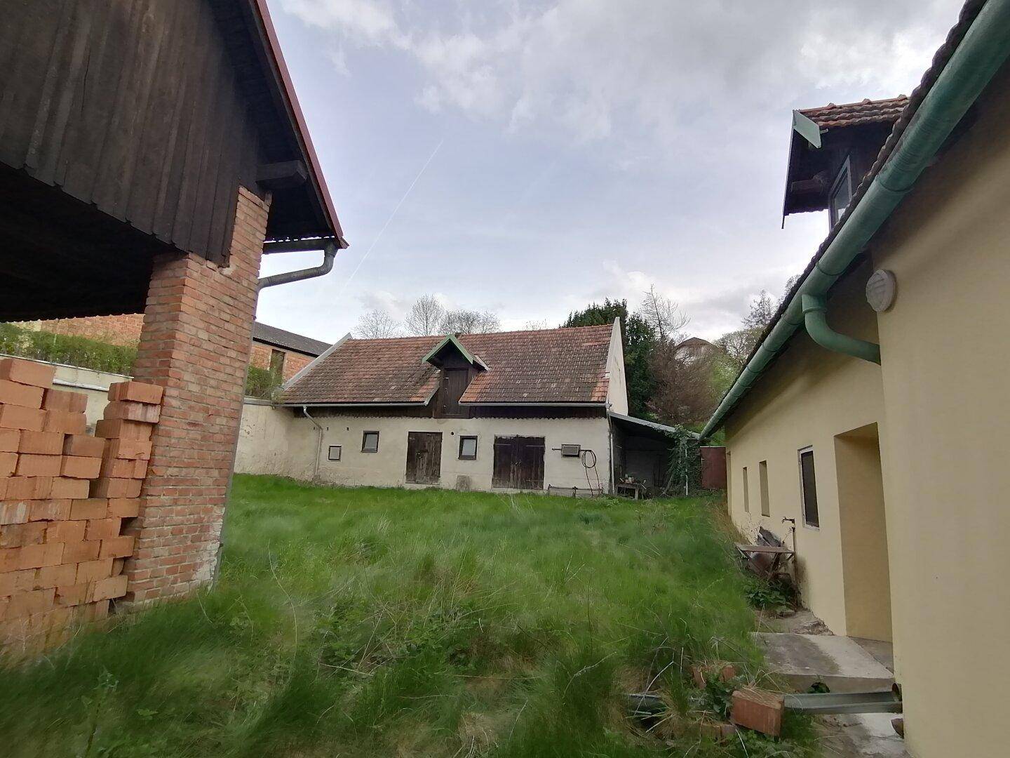 Blick auf ein älteres Gebäude mit Ziegeldach und einem verwilderten Gartenbereich im Vordergrund.