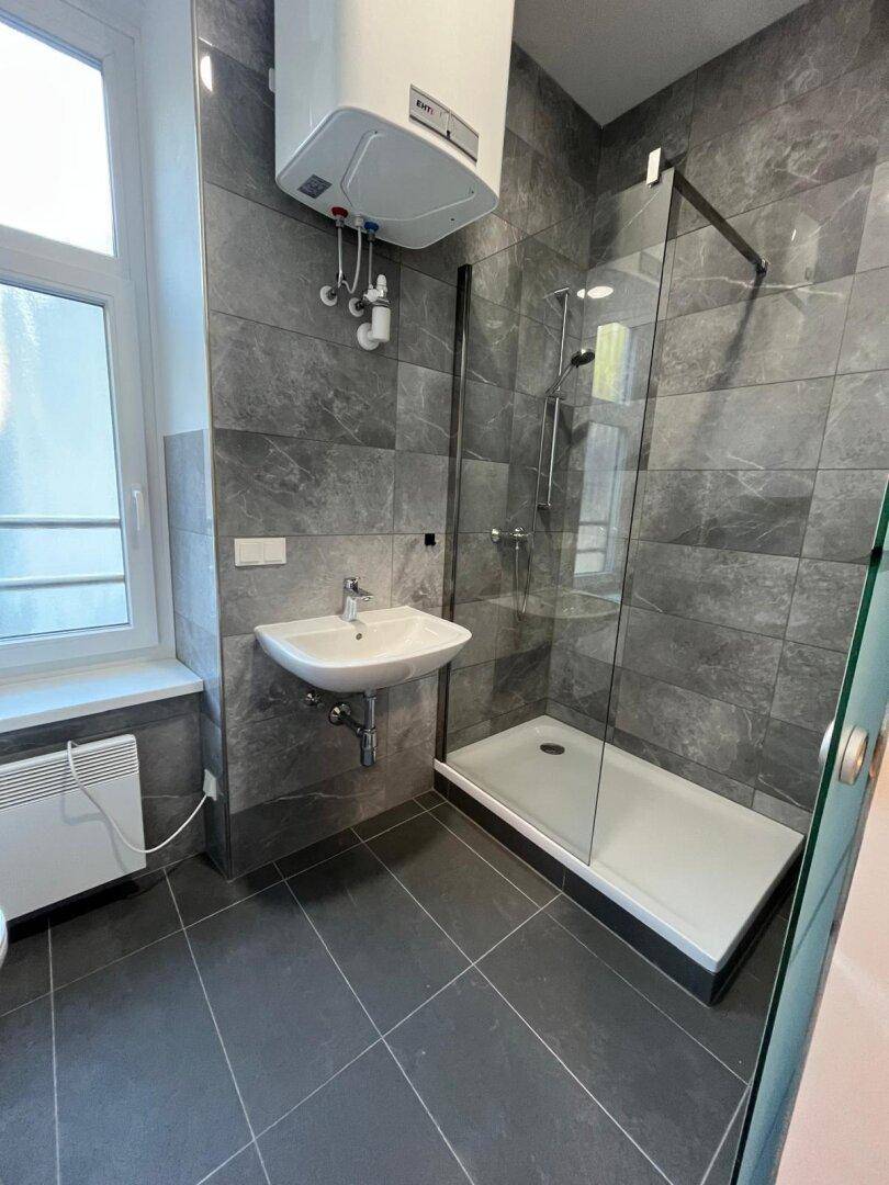 Modernes Badezimmer mit grauen Fliesen, Duschkabine und Fenster für Tageslicht.