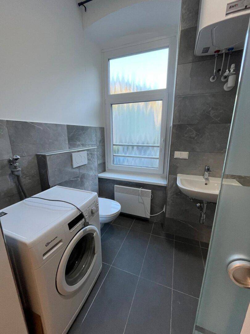 Funktionales Badezimmer mit grauen Fliesen, Toilette, Waschbecken und Waschmaschine.