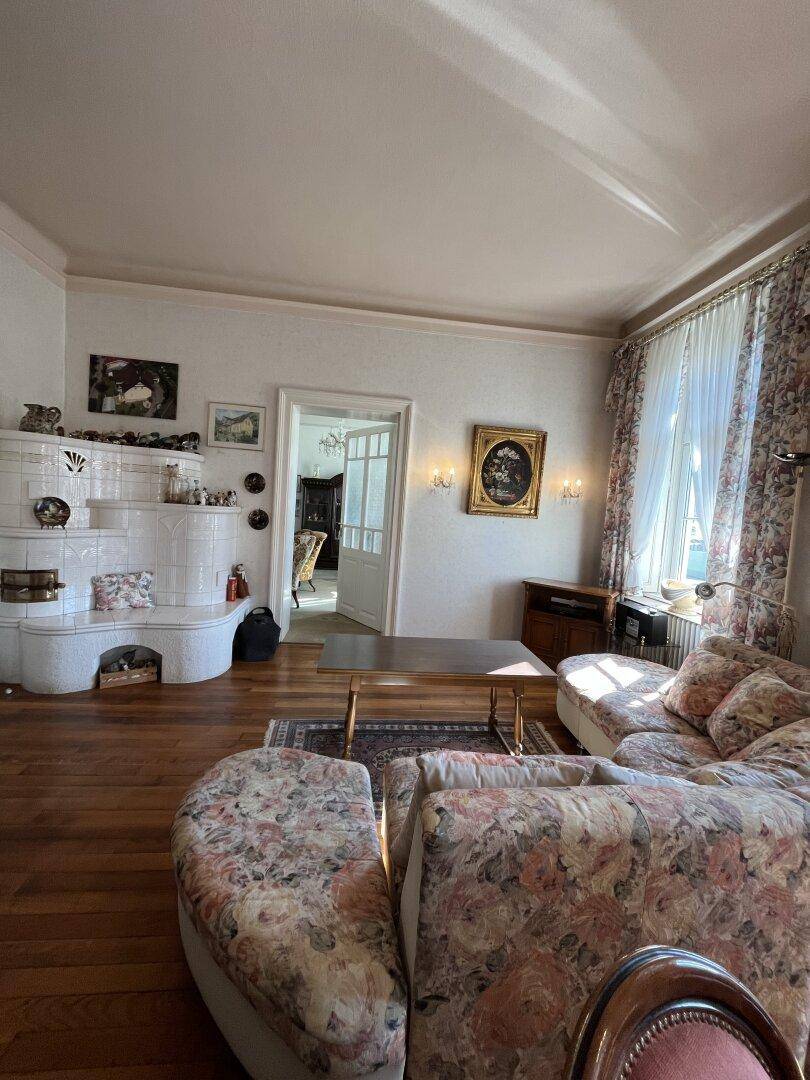 Geräumiges Wohnzimmer mit Kachelofen, floralen Vorhängen und Holzboden.