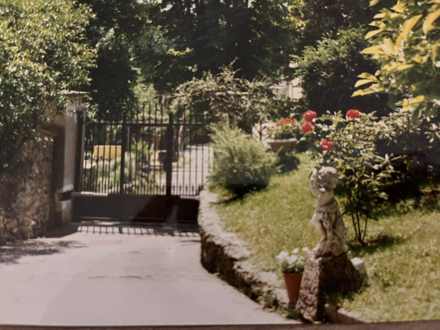 Garteneingang mit schmiedeeisernem Tor, üppiger Bepflanzung und Statue.