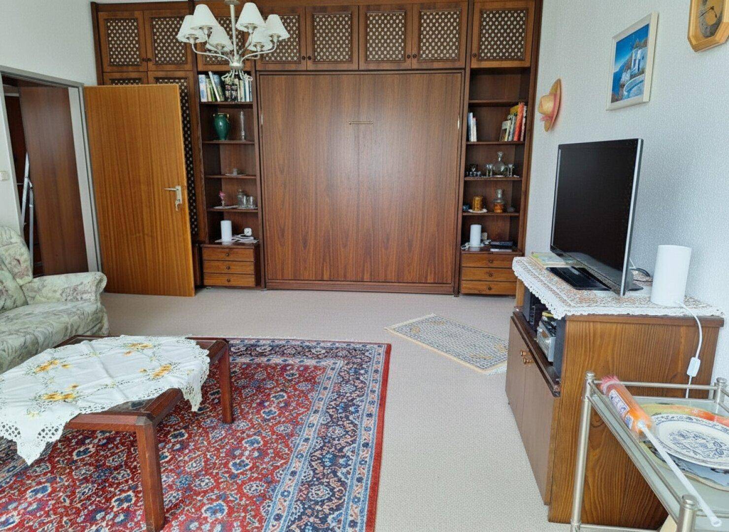 Wohnzimmer mit Schrankwand, integriertem Klappbett und traditionellem Teppich.