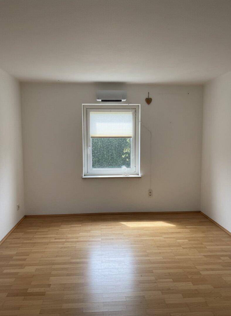 Leerer Raum mit hellem Laminatboden, Fenster und Klimaanlage.