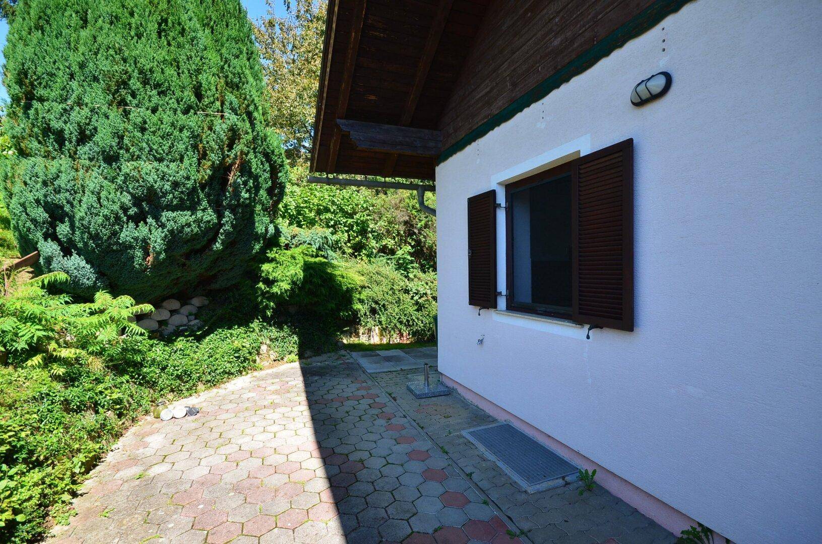Gepflasterter Weg entlang der Hausseite, umgeben von hohen Hecken und Bäumen.