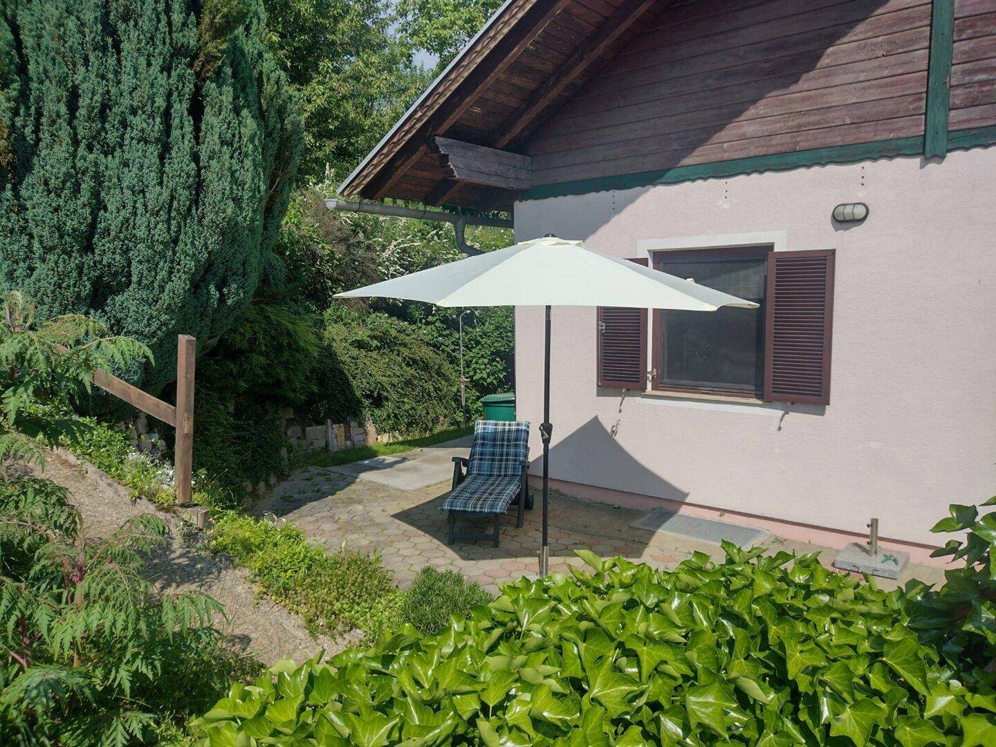 Gemütliche gepflasterte Terrasse mit Sonnenliege und Sonnenschirm, umgeben von üppigem Grün.