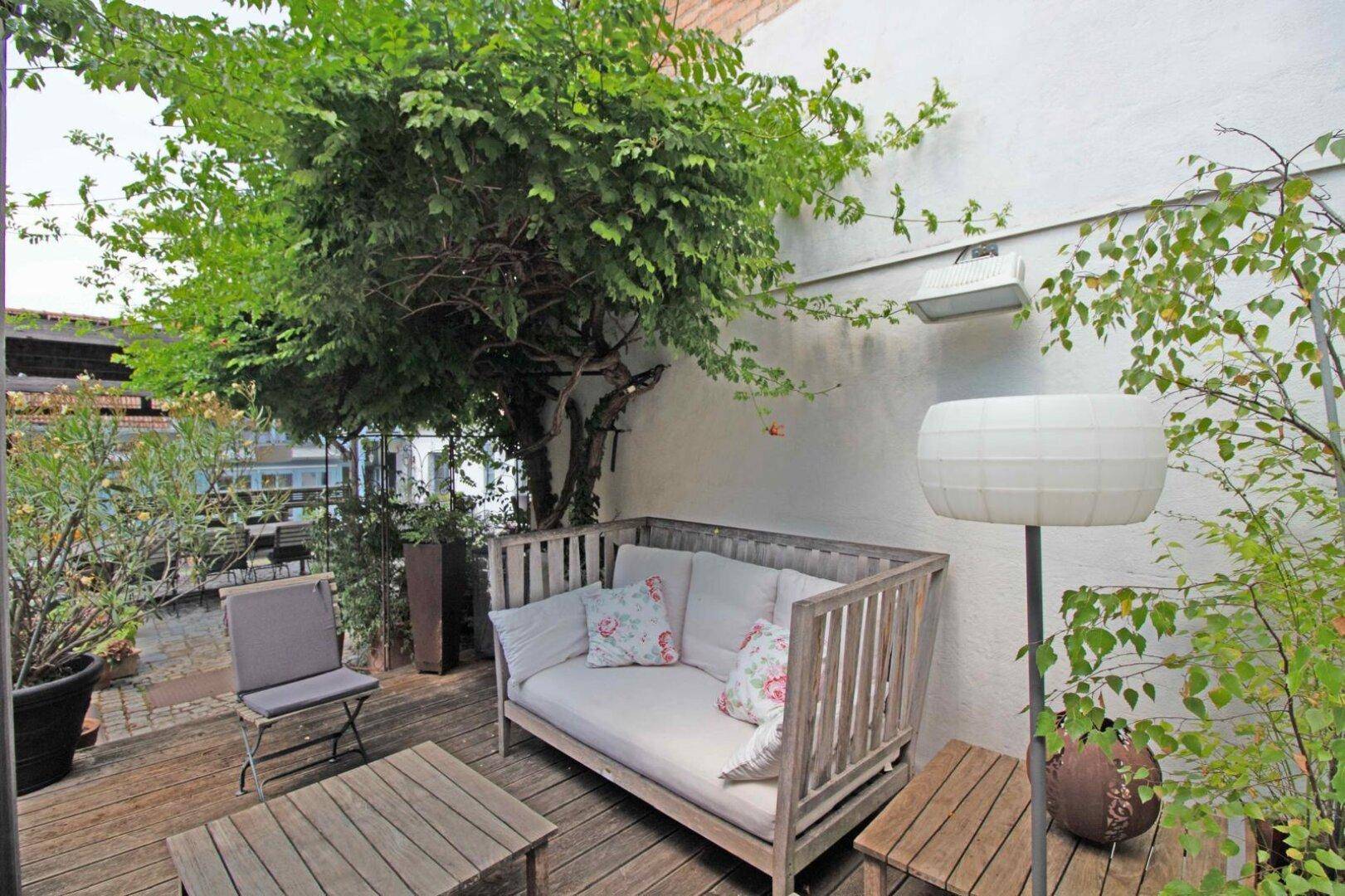 Gemütliche Terrasse mit Holzmöbeln, schattenspendendem Baum und weißer Hauswand.