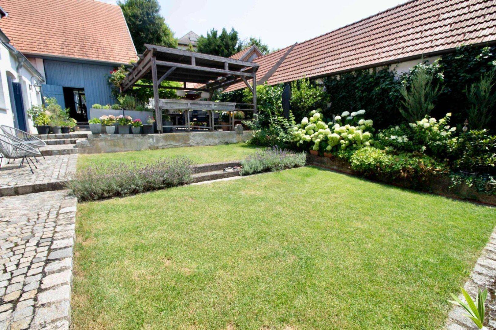 Gepflegter Garten mit Rasenfläche, Kopfsteinpflaster und einer Holzpergola im Hintergrund.