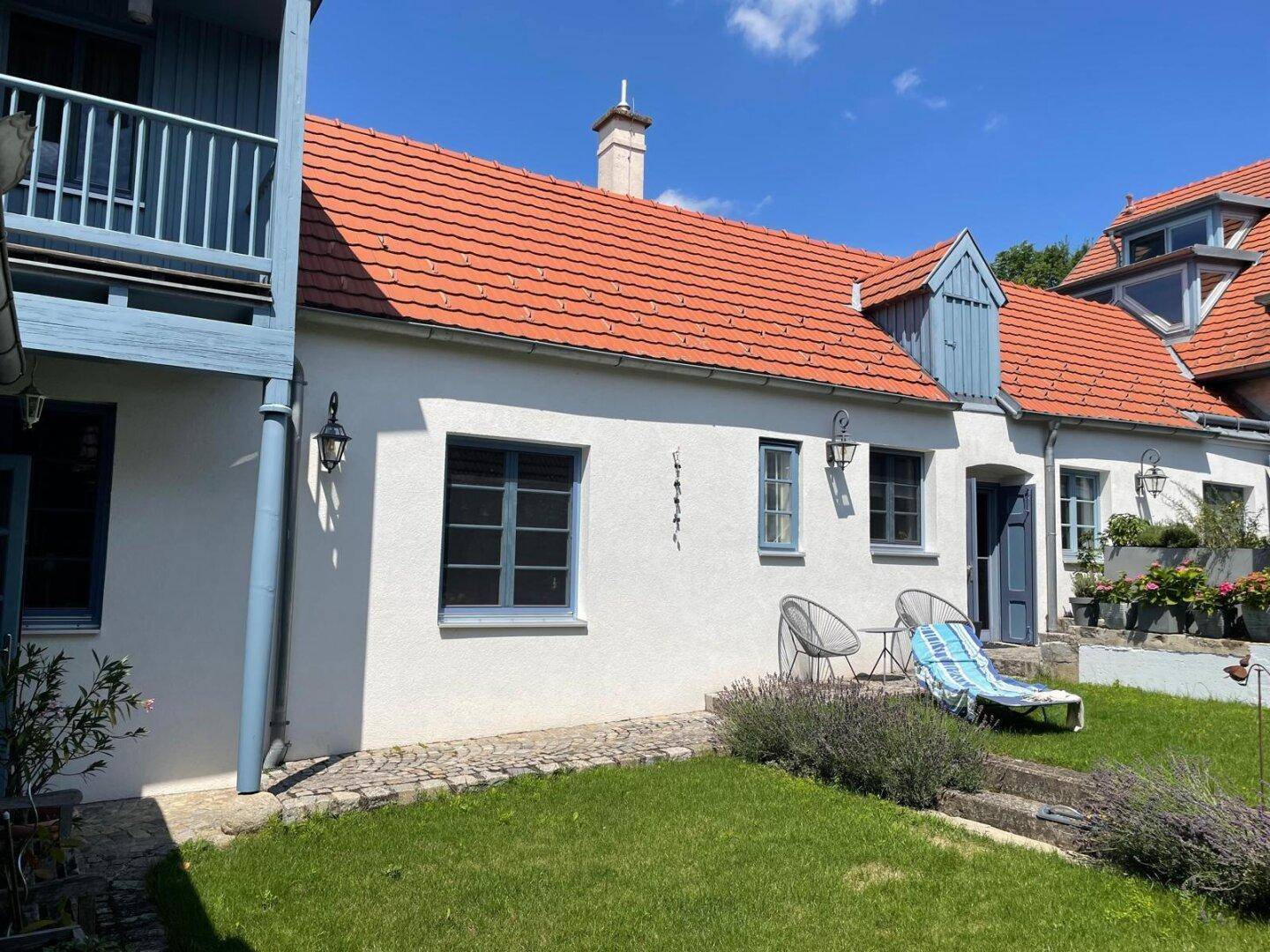 Weiße Hausfassade mit roten Dachziegeln, blauen Fensterläden und Rasenfläche.
