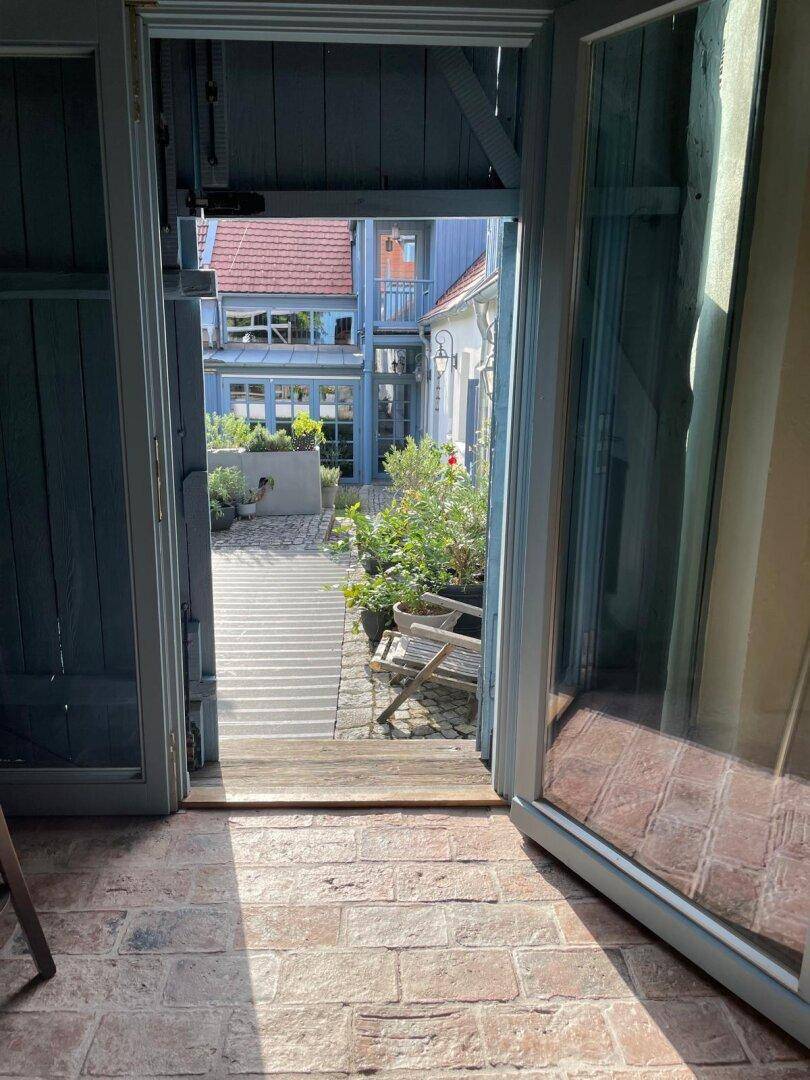Blick durch eine offene Tür auf den sonnigen Innenhof mit Pflanzen und Gebäuden.
