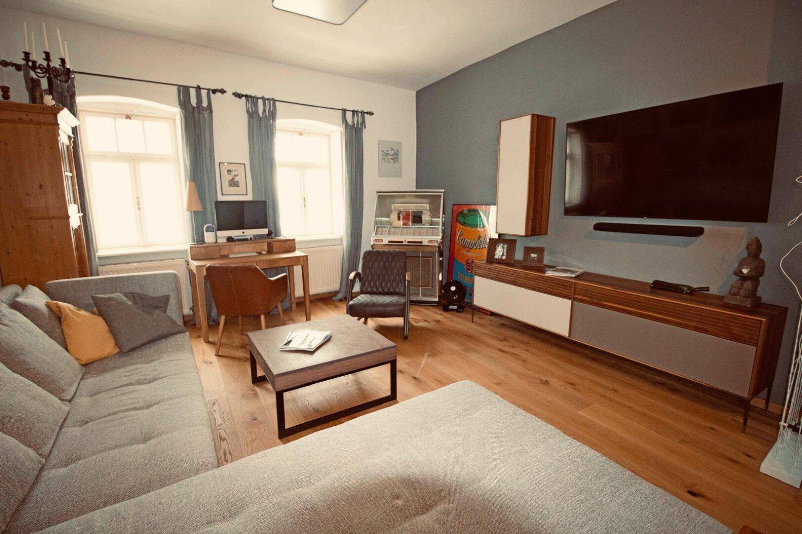 Gemütliches Wohnzimmer mit Sofa, Fernseher, Schreibtisch und Holzboden.