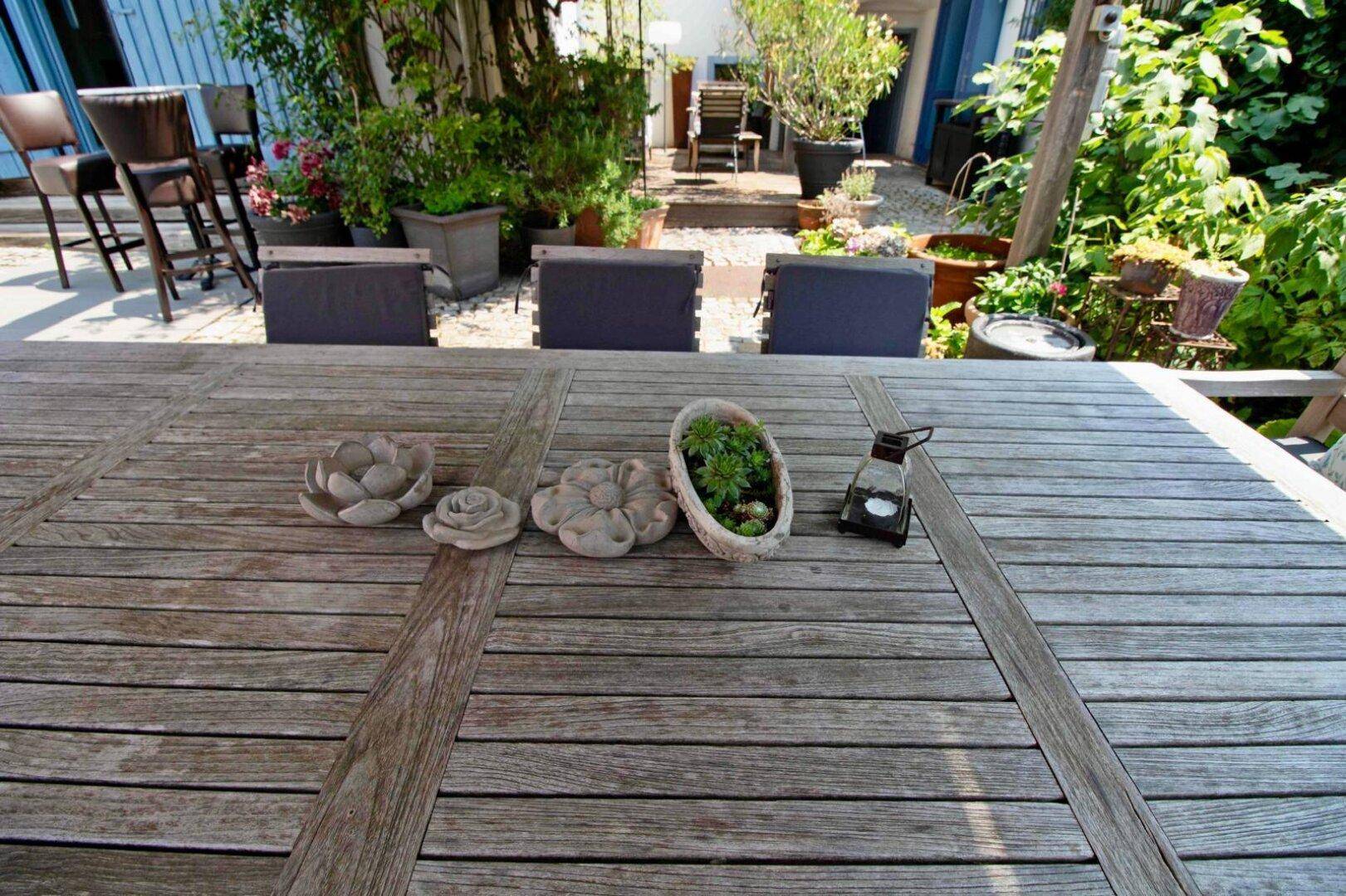 Großer Holztisch auf der Terrasse, umgeben von üppigem Grün und Topfpflanzen im Garten.