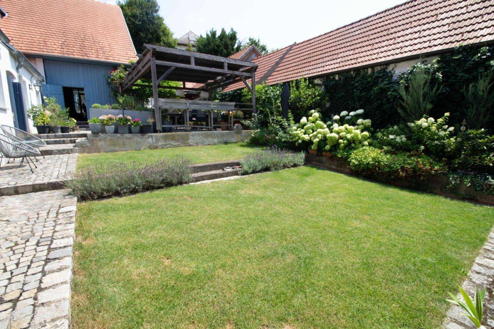 Gepflegter Garten mit Rasenfläche, Kopfsteinpflaster und einer Holzpergola im Hintergrund.