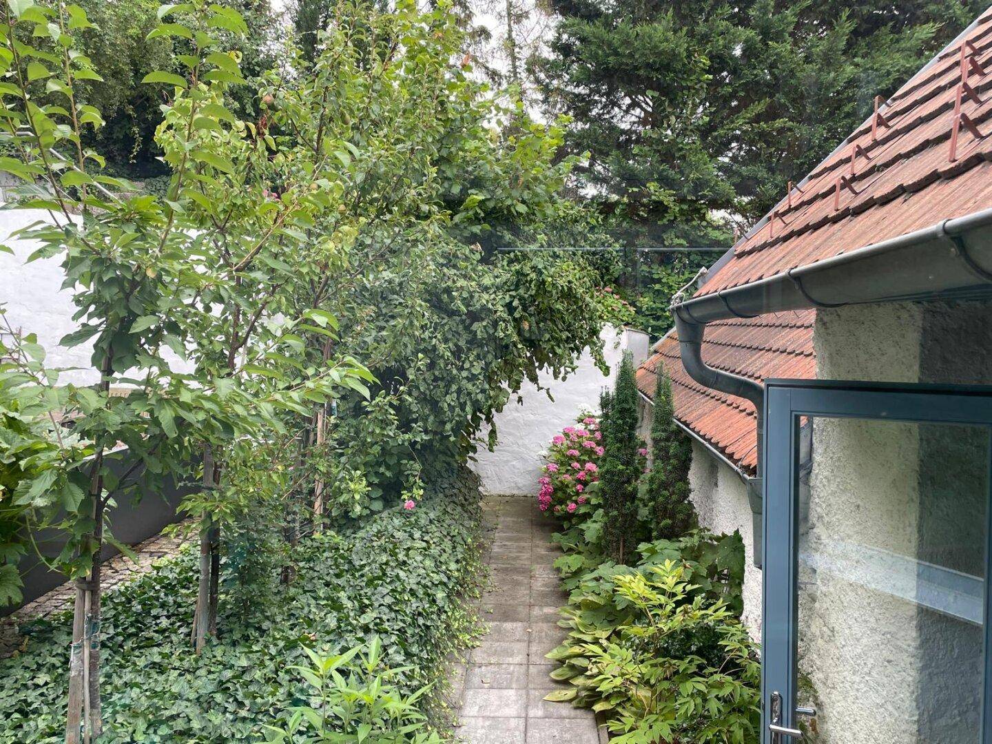 Schmaler Gartenweg zwischen üppigem Grün und einem Haus mit traditionellem Ziegeldach.