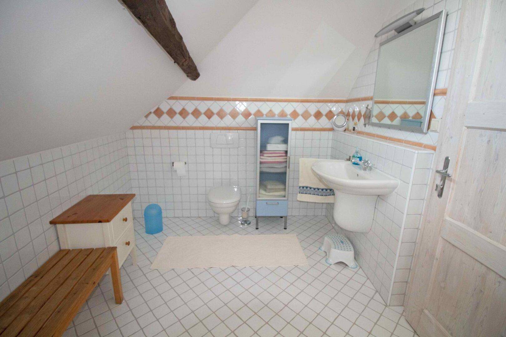 Helles Badezimmer im Dachgeschoss mit weißen Fliesen, Dachschräge und sichtbarem Holzbalken.