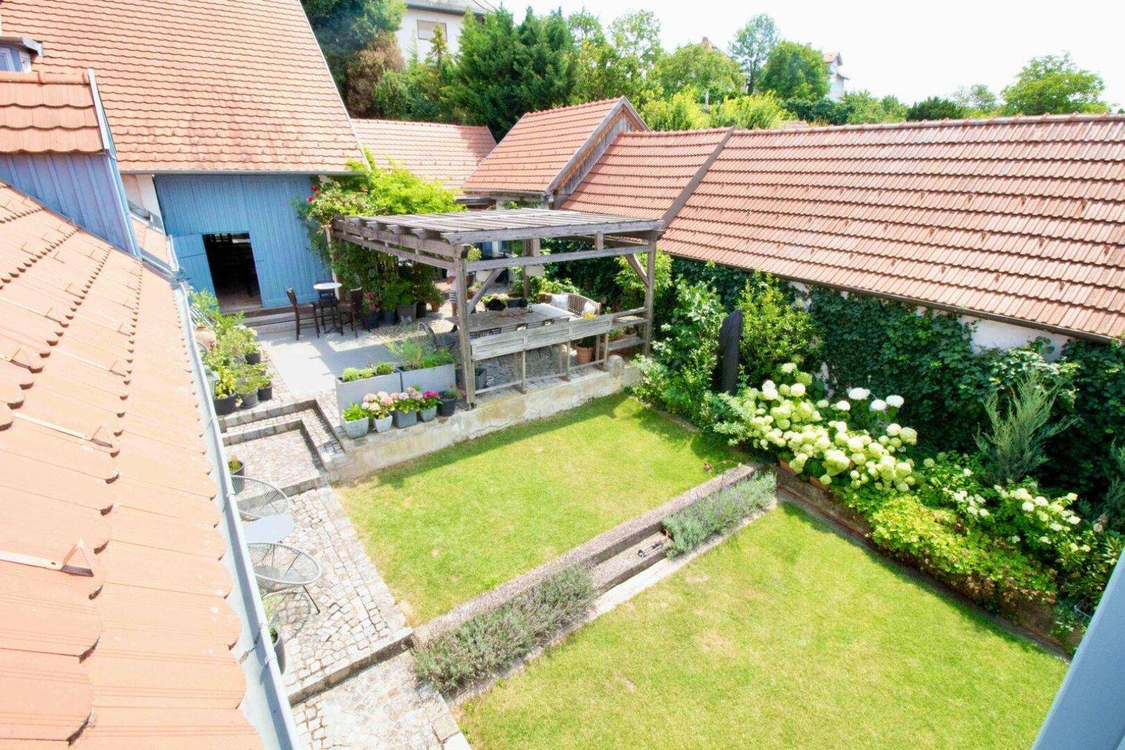 Vogelperspektive auf den Gartenbereich mit Rasen, Pergola und angrenzenden Gebäuden mit Ziegeldächern.