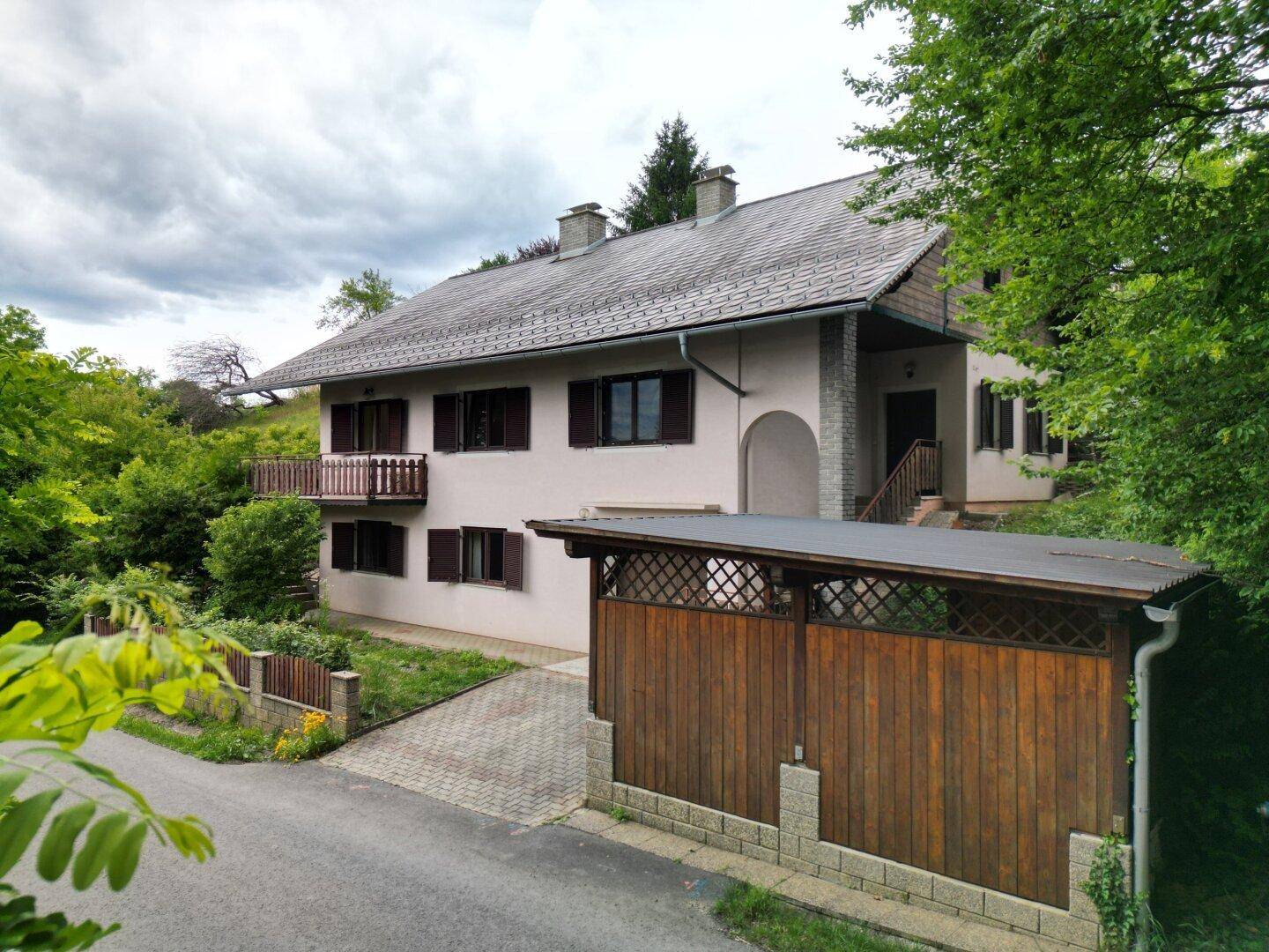 Wohnhaus mit Balkon und Carport, umgeben von Bäumen und einer gepflasterten Zufahrt zum Grundstück.