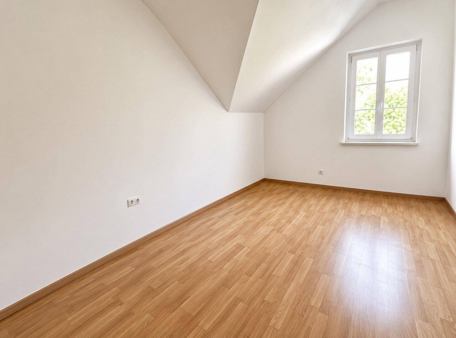 Leeres Zimmer im Dachgeschoss mit schräger Decke, Laminatboden und großem Fenster.