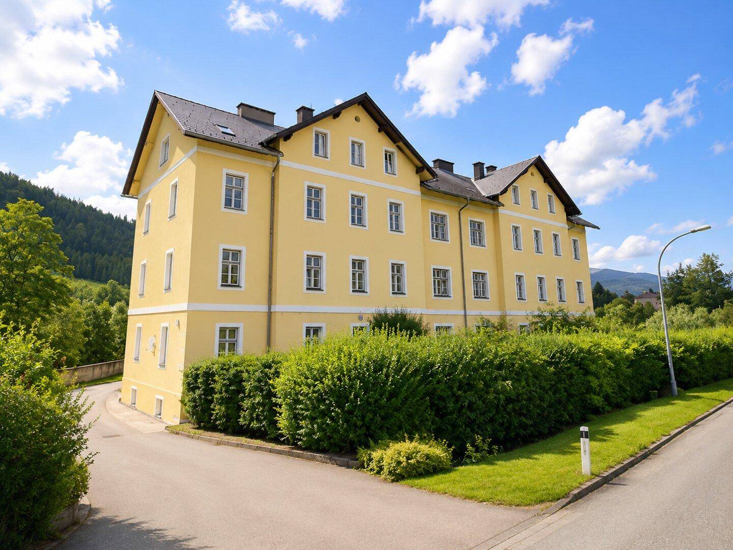Großes, gelbes Gebäude mit traditioneller Architektur, umgeben von grüner Landschaft und Bäumen.