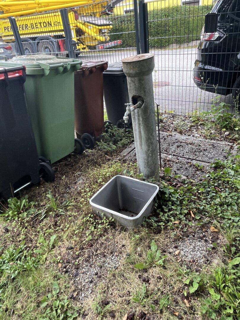 Außenbereich mit Mülltonnen und einem Wasserhahn, umgeben von Gras und einem Zaun.