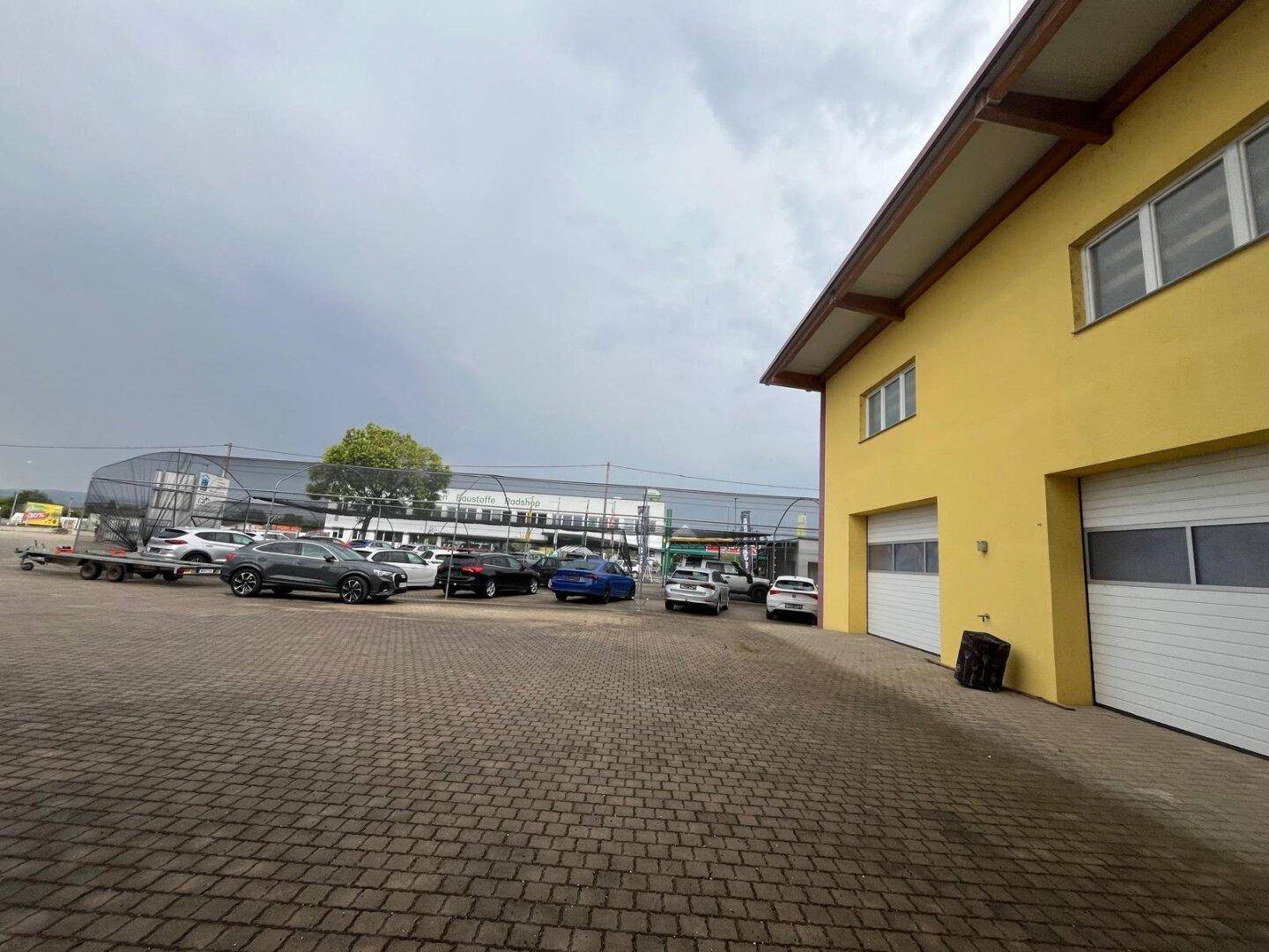 Großer gepflasterter Parkplatz mit mehreren Fahrzeugen neben dem gelben Geschäftsgebäude mit Garagentoren.