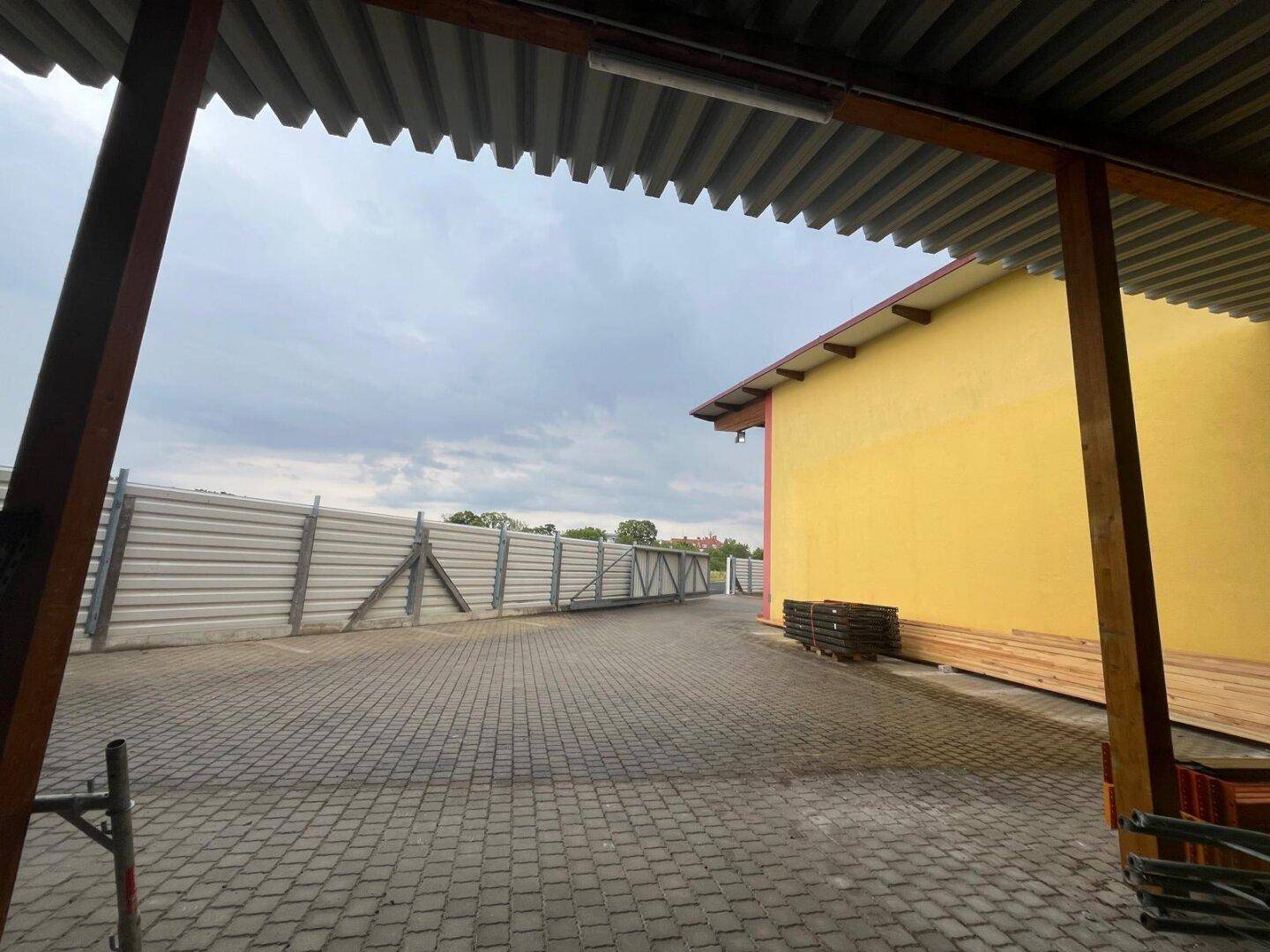 Blick von einem überdachten Bereich auf einen gepflasterten Hof und einen hohen Metallzaun.