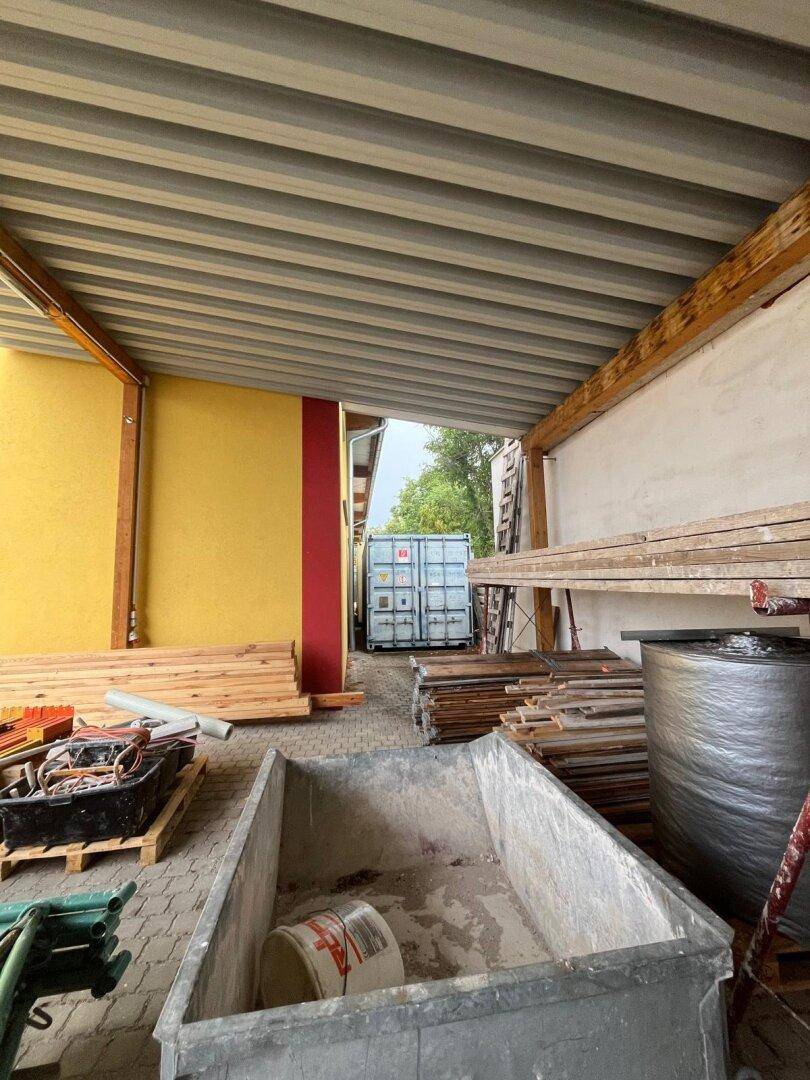 Überdachter Lagerbereich mit Holzregalen, Baumaterialien und einem Container im Hintergrund.