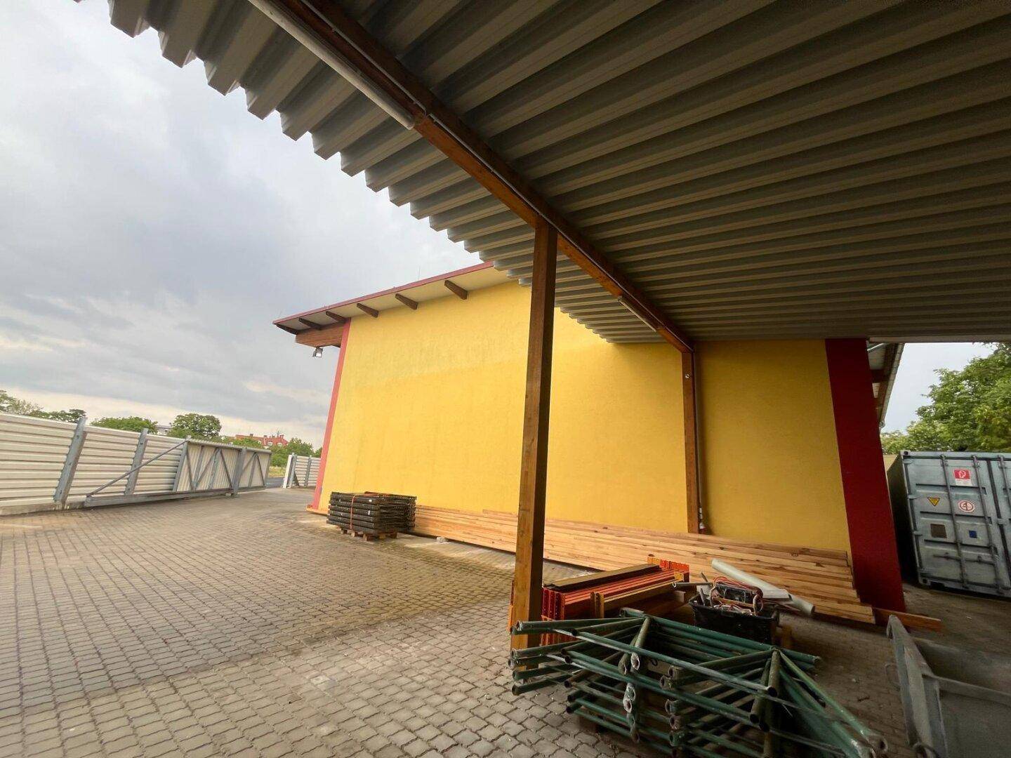 Überdachter Außenbereich mit gepflastertem Boden, Metallzaun und gelagerten Baumaterialien.
