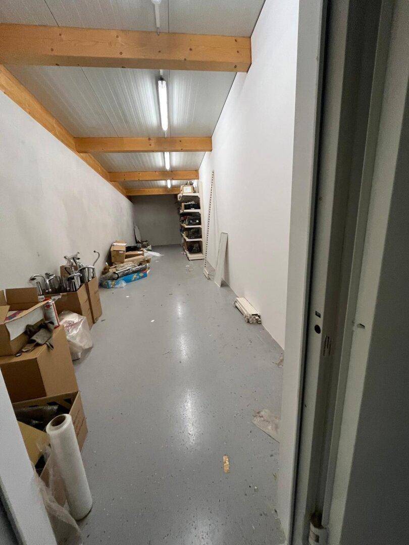 Langer, schmaler Lagerraum mit Holzbalken, Betonboden und Regalen voller Gegenstände.