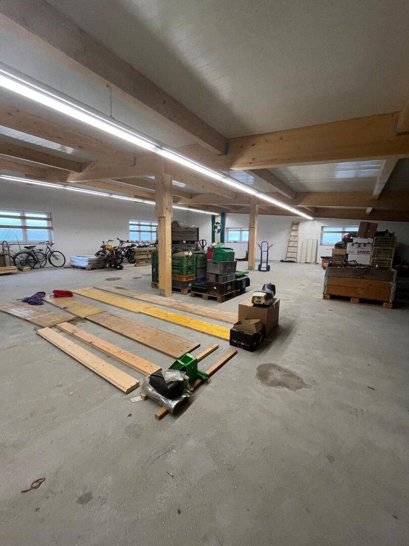 Geräumige Lagerhalle mit Holzbalken, Betonboden und großen Fenstern, gefüllt mit Materialien.