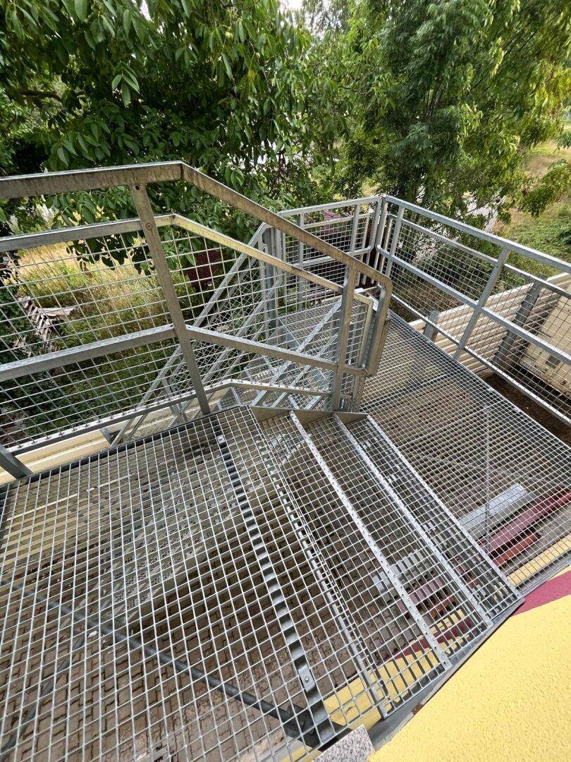 Außenliegende Metalltreppe mit Gitterroststufen und Geländer, umgeben von Bäumen.