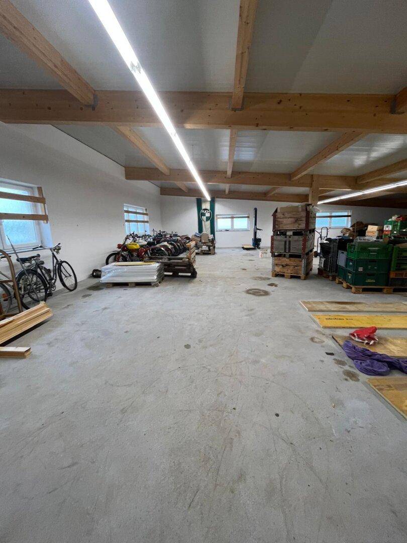 Geräumige Lagerhalle mit Holzbalken, Betonboden und großen Fenstern, gefüllt mit Materialien.