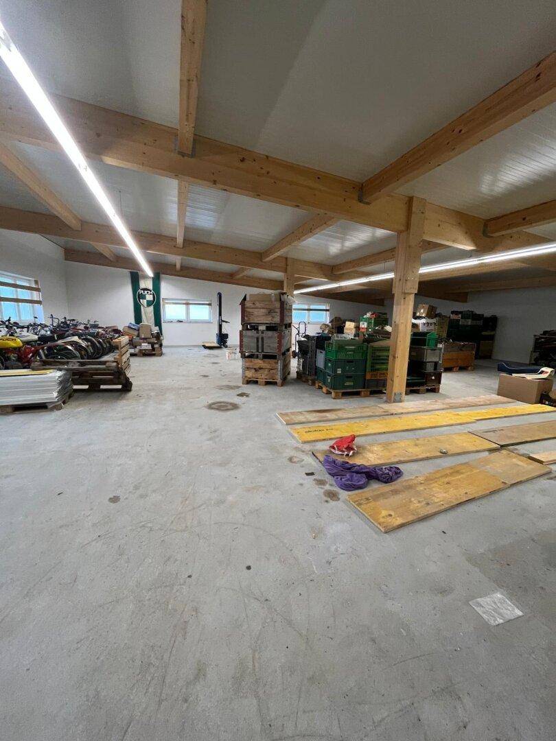 Geräumige Lagerhalle mit Holzbalken, Betonboden und großen Fenstern, gefüllt mit Materialien.