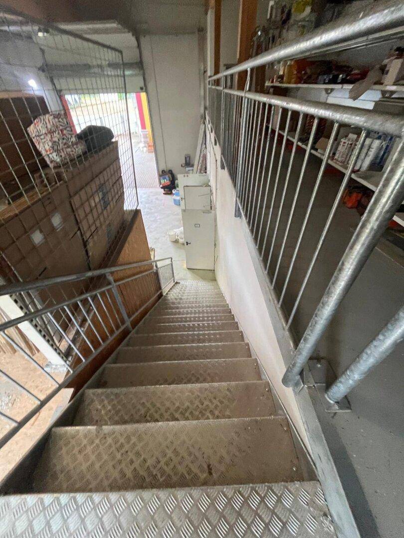 Metalltreppe führt in einen Lagerbereich mit Regalen und verschiedenen Materialien, gut beleuchtet.