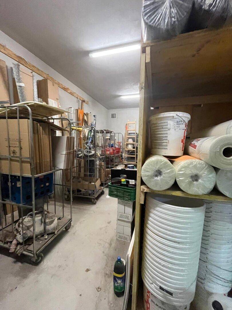 Lagerraum mit Regalen, Rollwagen und Baumaterialien, beleuchtet von Deckenleuchten und hellen Wänden.