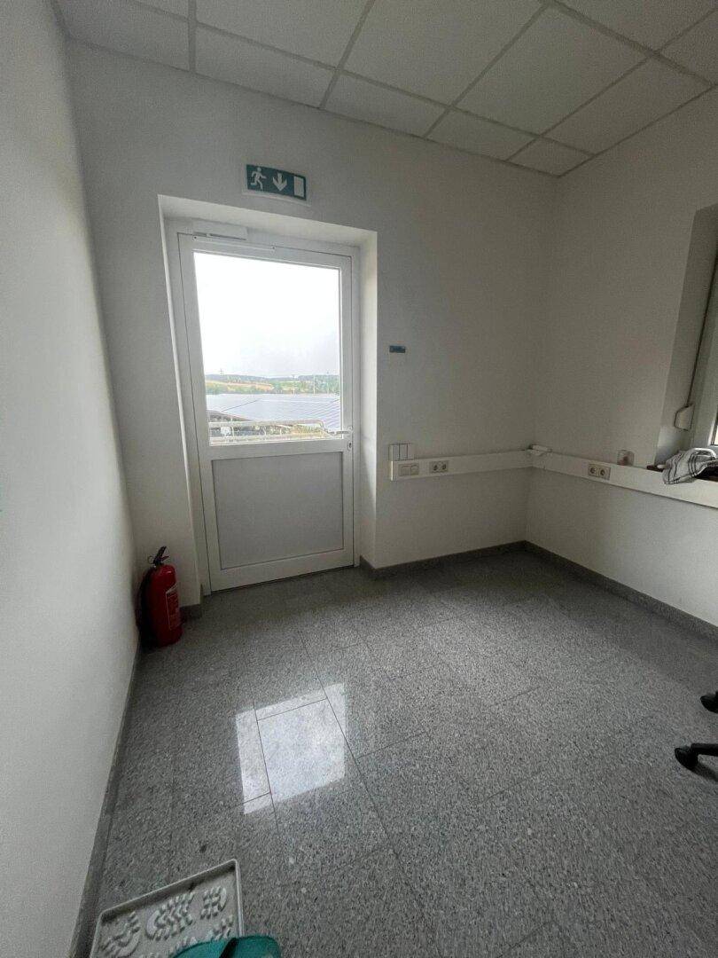 Leeres Büro mit grauem Fliesenboden, weiß gestrichenen Wänden und einer Tür mit Blick nach draußen.